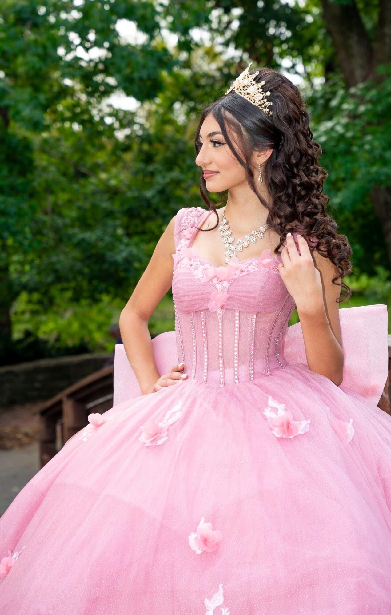 Young woman in pink dress and tiara outdoors, smiling, with trees in background.