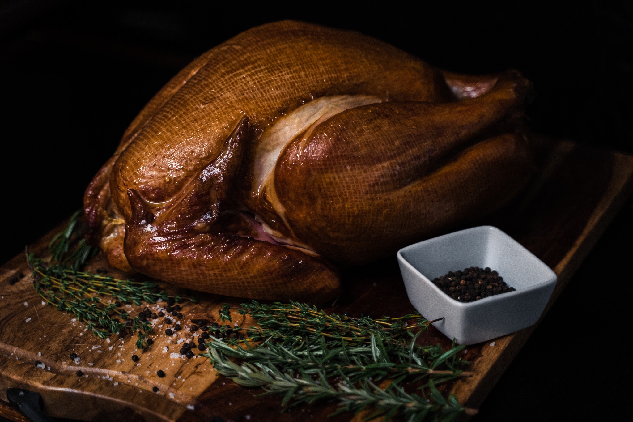 Roast turkey on a wooden cutting board with herbs and a small bowl of black pepper, dark background, close-up.