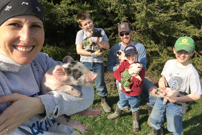Group of children and an adult outdoors with a woman holding a piglet, all smiling, in a grassy area with trees.