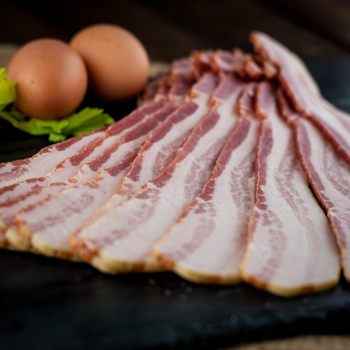 Raw bacon slices arranged on a dark surface with eggs and lettuce in the background.