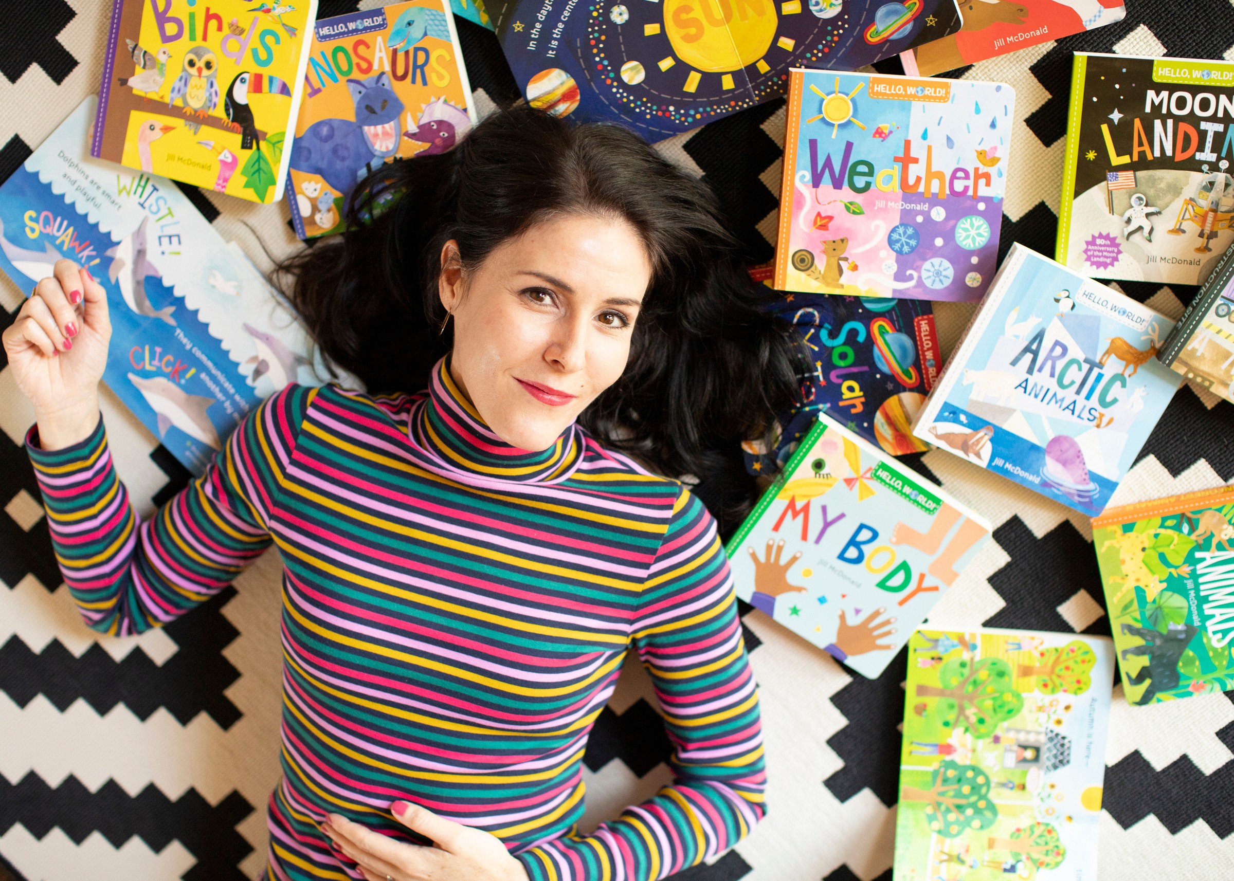 Woman lying on black and white zigzag patterned surface surrounded by colorful children's books, wearing a striped long-sleeve shirt, looking at the camera.
