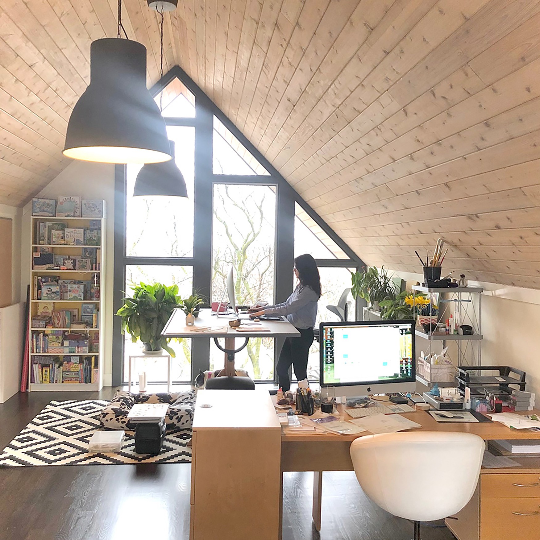 Person sitting at a desk near large windows with plants, bookshelf, and computer in a room with wooden ceiling.