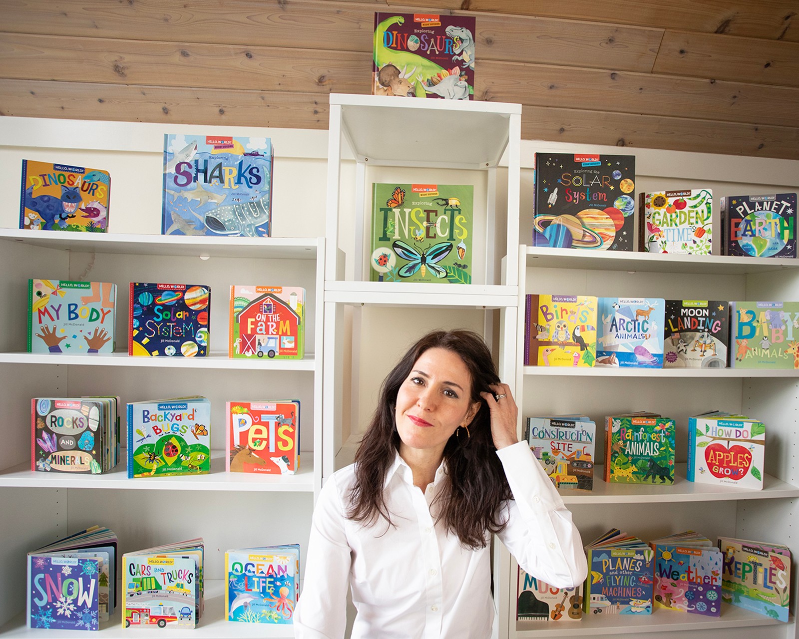 Woman with dark hair in white shirt standing in front of shelves filled with colorful children's books and games.