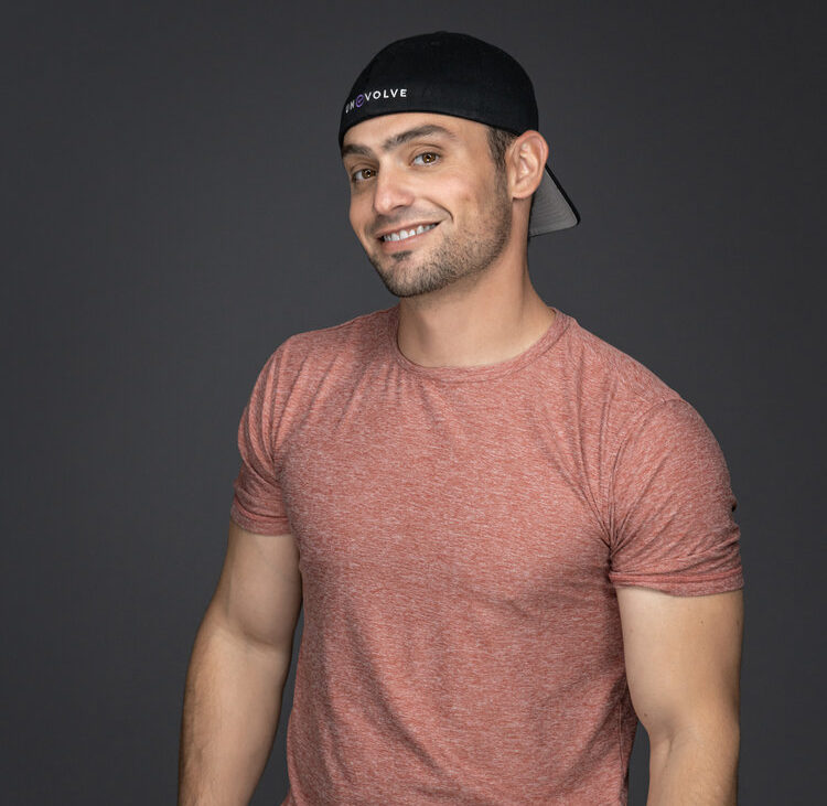 Young man with a cap smiling, wearing a red t-shirt, against a dark background.