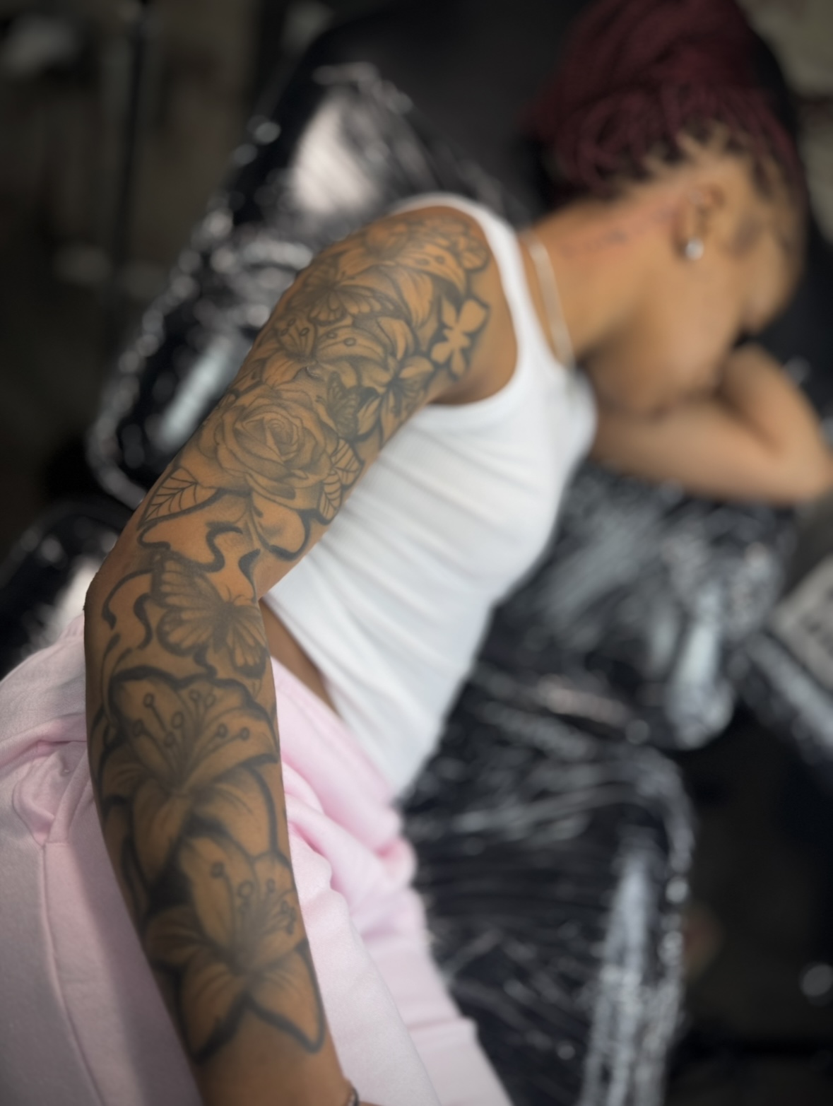 Person with a large tattoo sleeve on their arm, wearing a white tank top and pink pants, leaning forward.