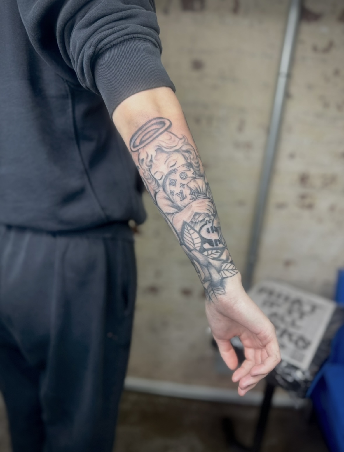 Person's arm with a detailed black and gray tattoo sleeve, wearing a dark long-sleeve shirt, against a brick wall background.