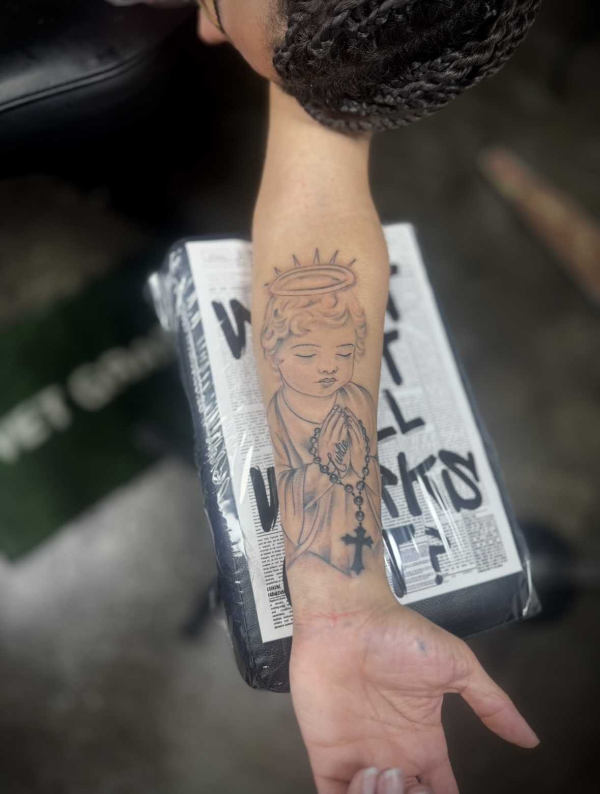 Person's forearm with a tattoo of a child's face, hands in prayer, and a cross, resting on a box with text.