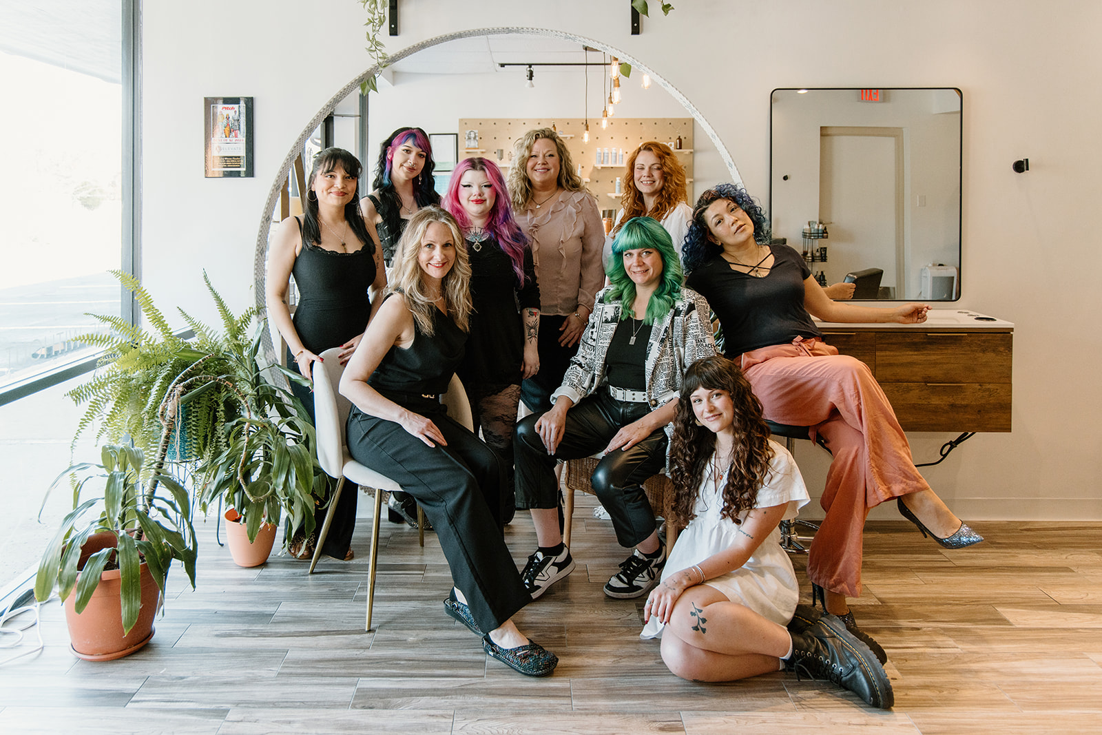 Group of nine women posing indoors, some sitting and some standing, with plants and a mirror in the background.