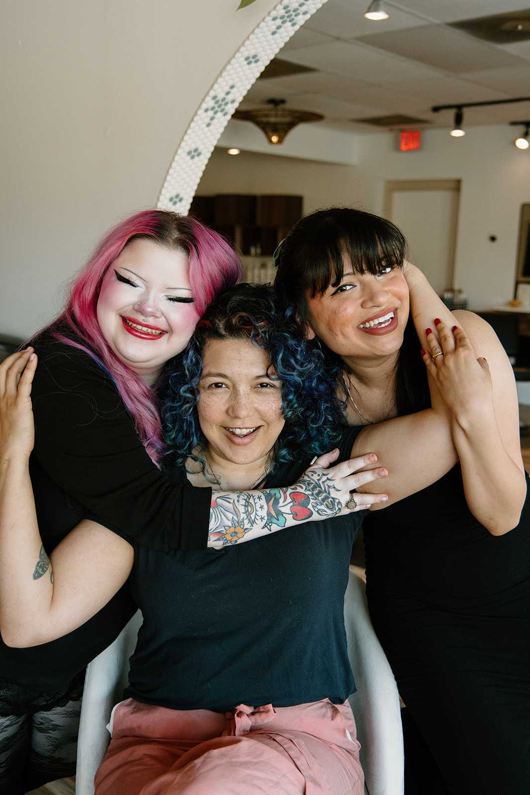 Three women smiling and hugging indoors, one with pink hair, one with dark curly hair, and one with straight dark hair.