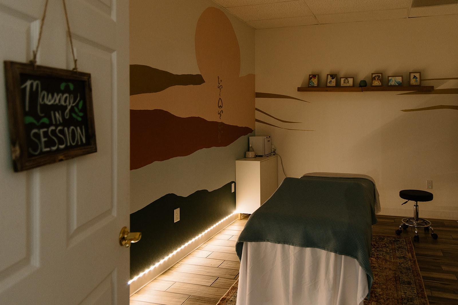Massage table in a room with wall art, a shelf with framed photos, and a stool, illuminated by warm lighting.