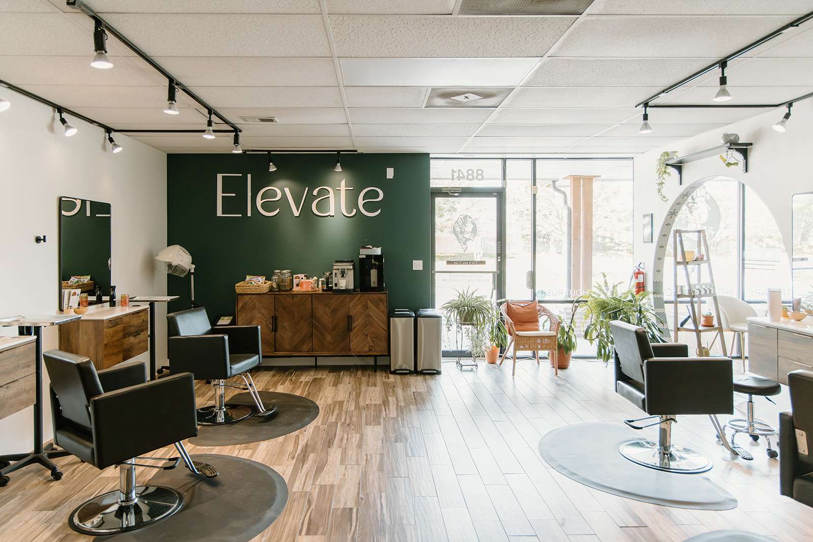 Salon with chairs, a green wall with 'Elevate' sign, and large windows letting in natural light.