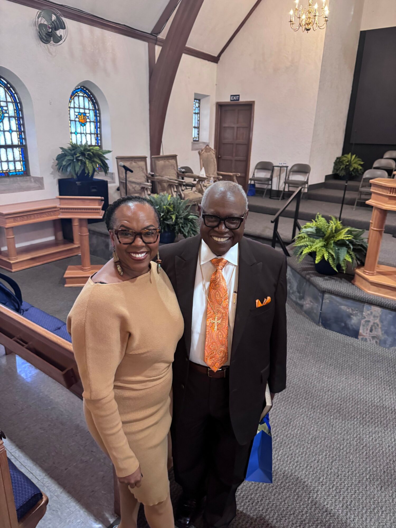 Two people standing inside a church, one in a black suit and orange tie, the other in a beige outfit, smiling.