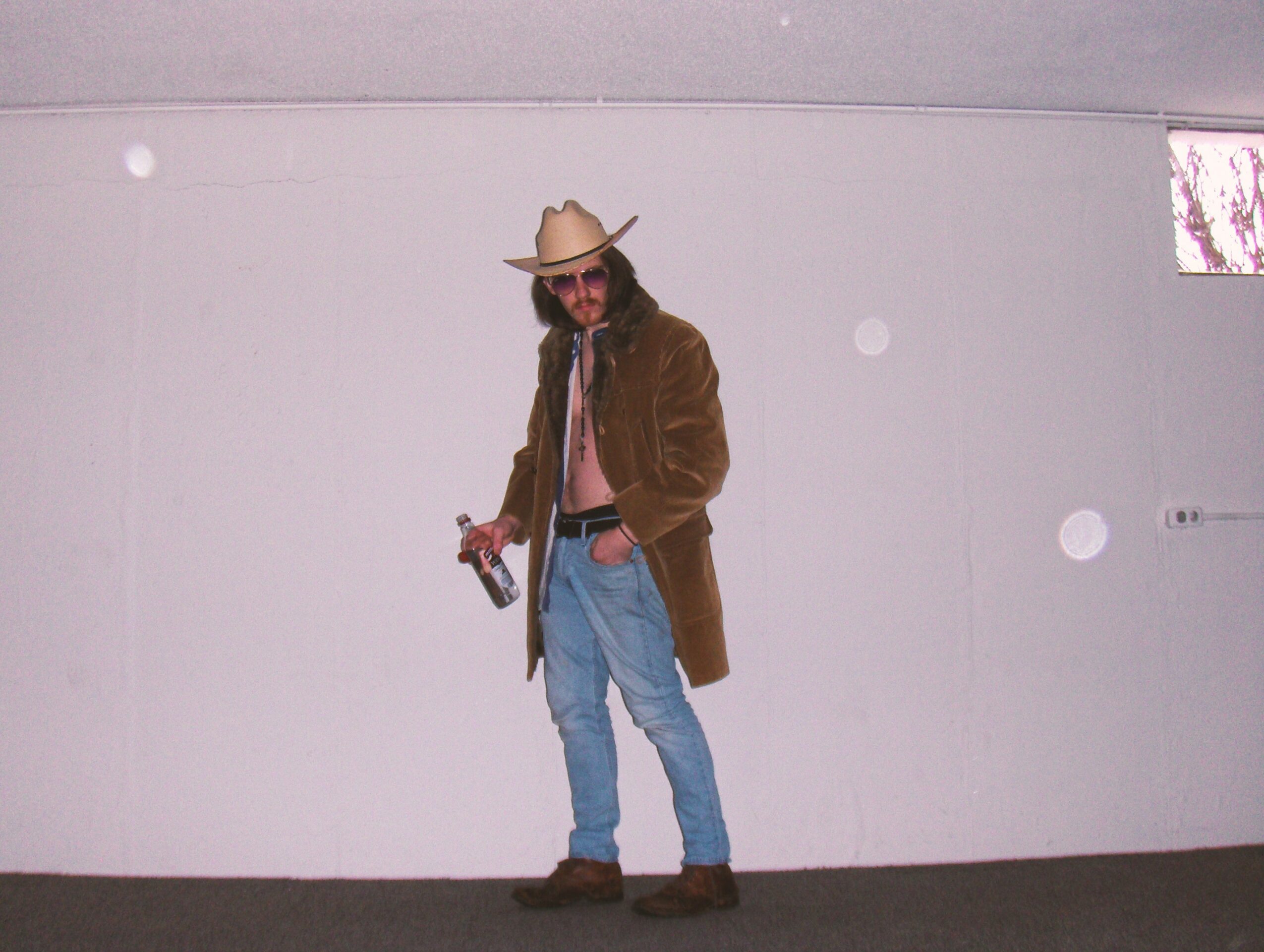 Person wearing a cowboy hat, sunglasses, brown jacket, and jeans, holding a bottle, standing against a plain wall.