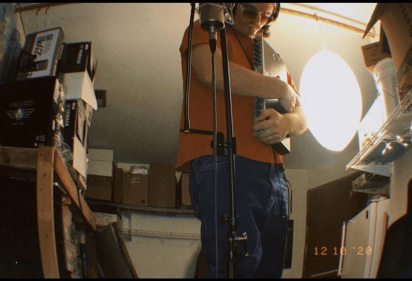 Person taking a mirror selfie in a cluttered room with shelves and boxes, holding a phone, wearing glasses and an orange top.