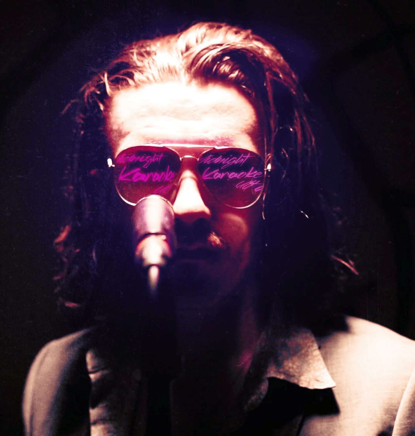 Person with long hair wearing sunglasses, speaking into a microphone, dark background