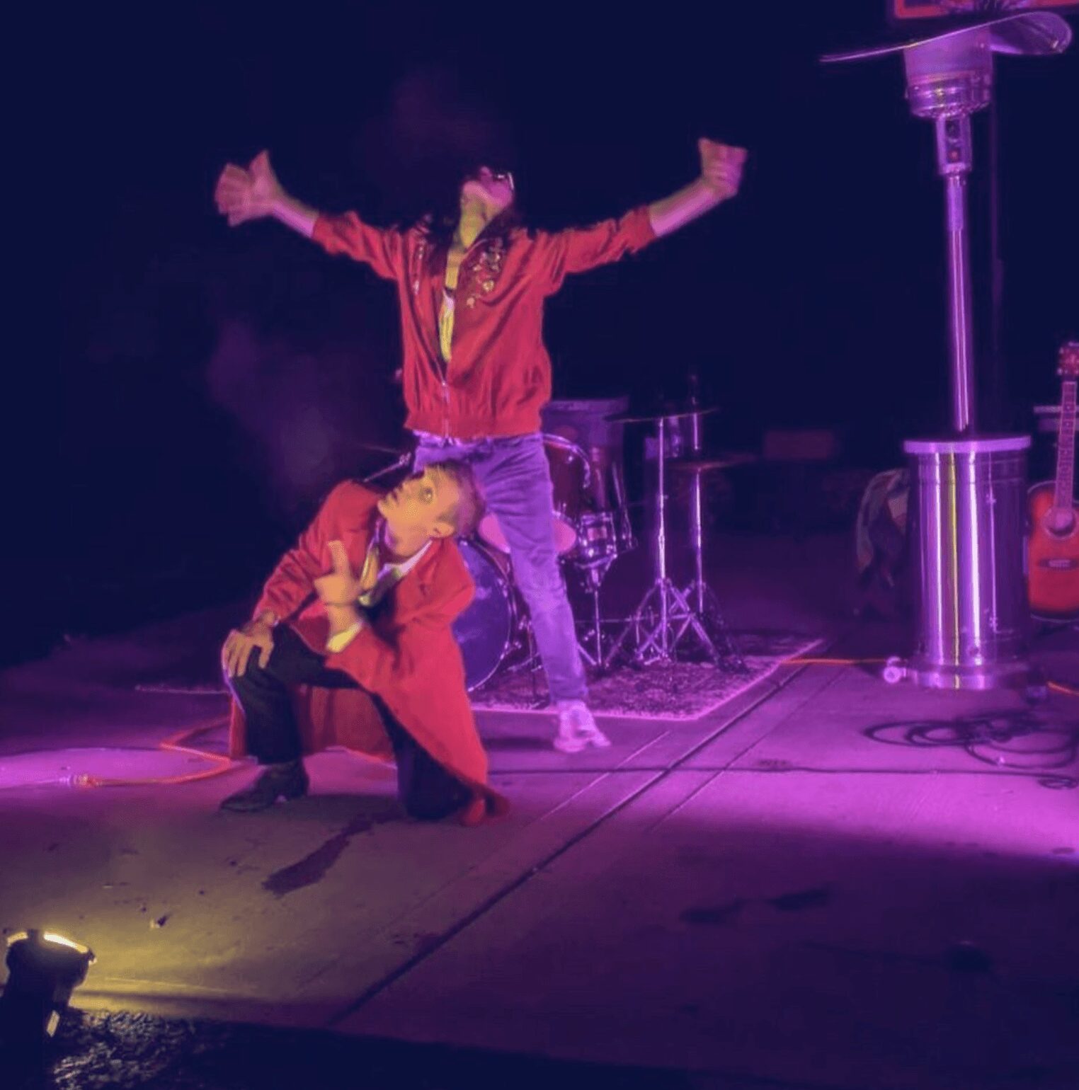 Two performers on stage, one standing with arms outstretched, the other kneeling with hand on face, stage with musical instruments nearby.