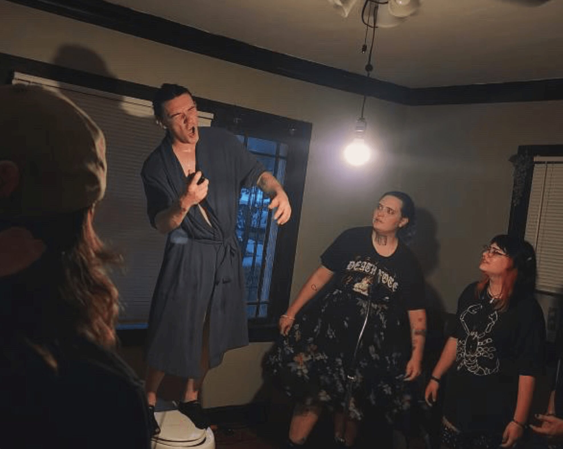 Person standing on a platform speaking to a group in a dimly lit room with a window and hanging light bulb.