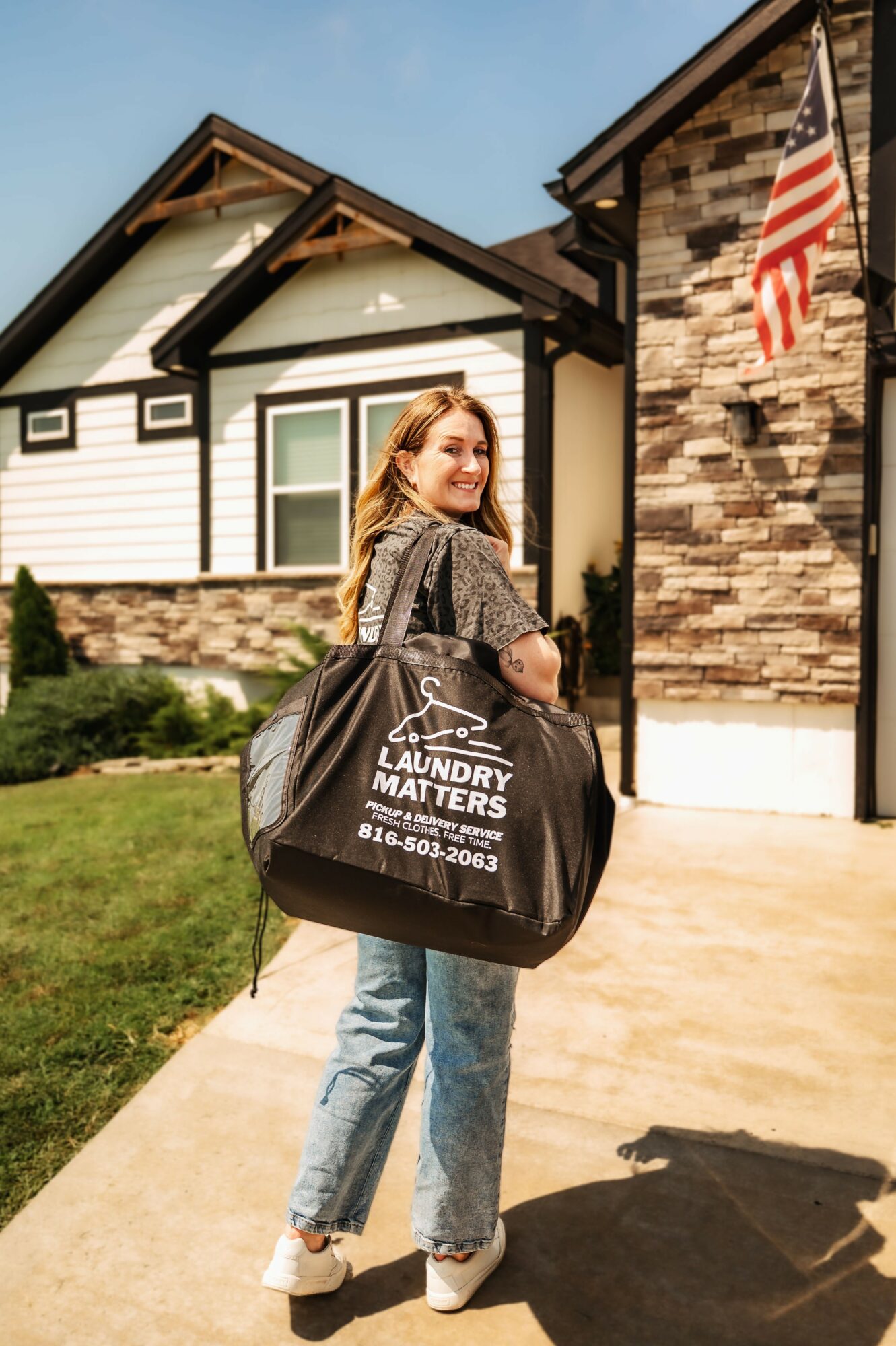 Young woman with a laundry bag standing outside a house, smiling, with American flag and house in background.