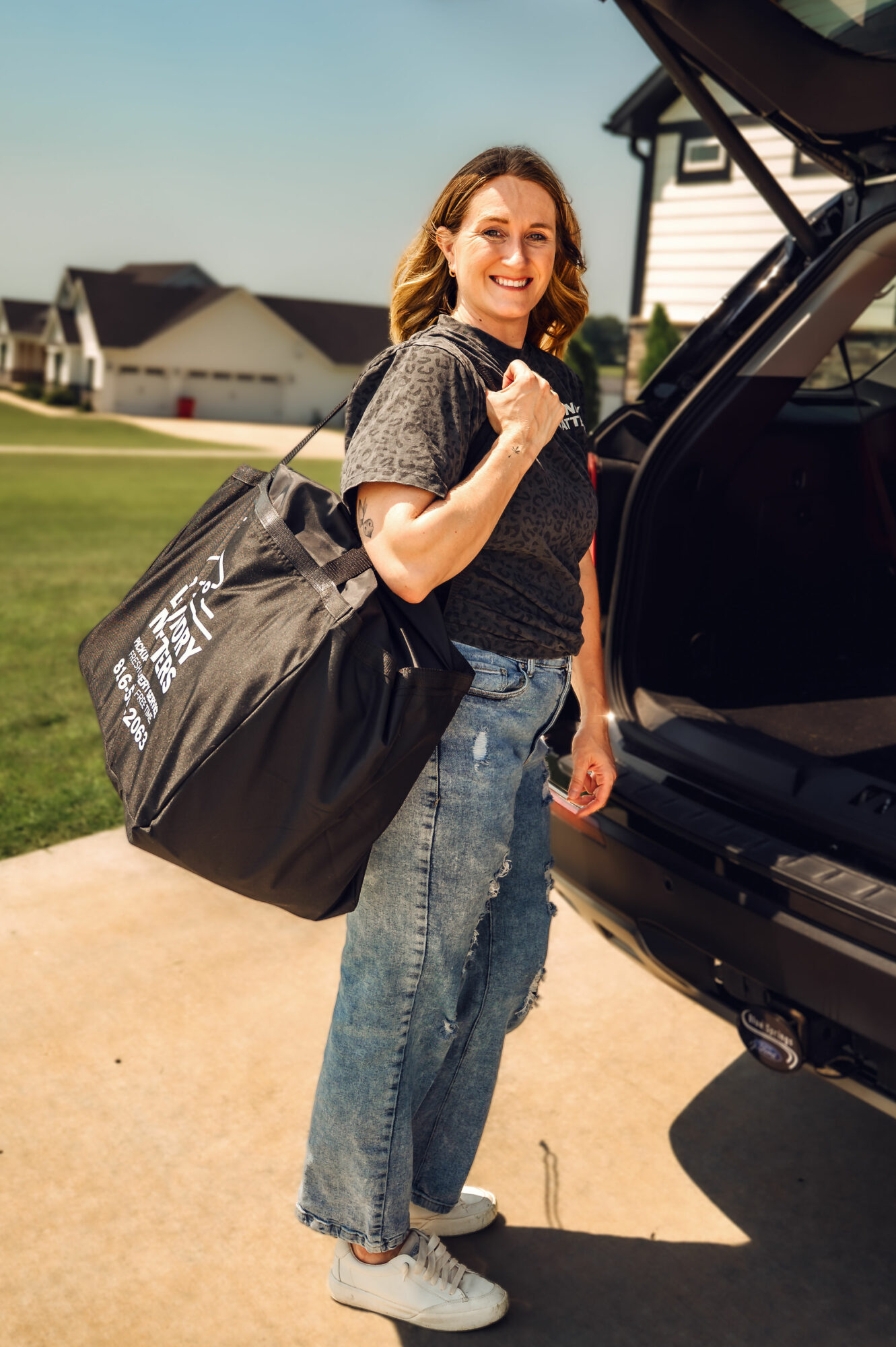Woman with a large black bag standing outdoors near a car trunk, smiling, in a residential neighborhood.