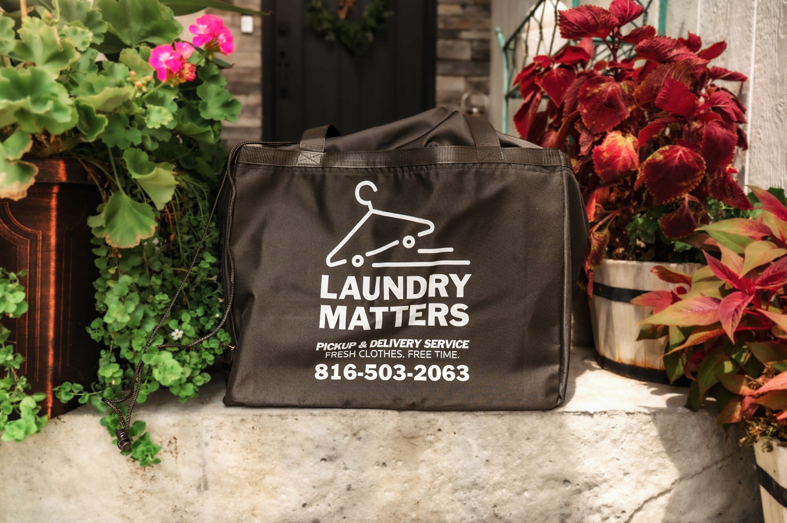 Black laundry bag with white logo and text, surrounded by potted pink and red flowers on a stone surface.