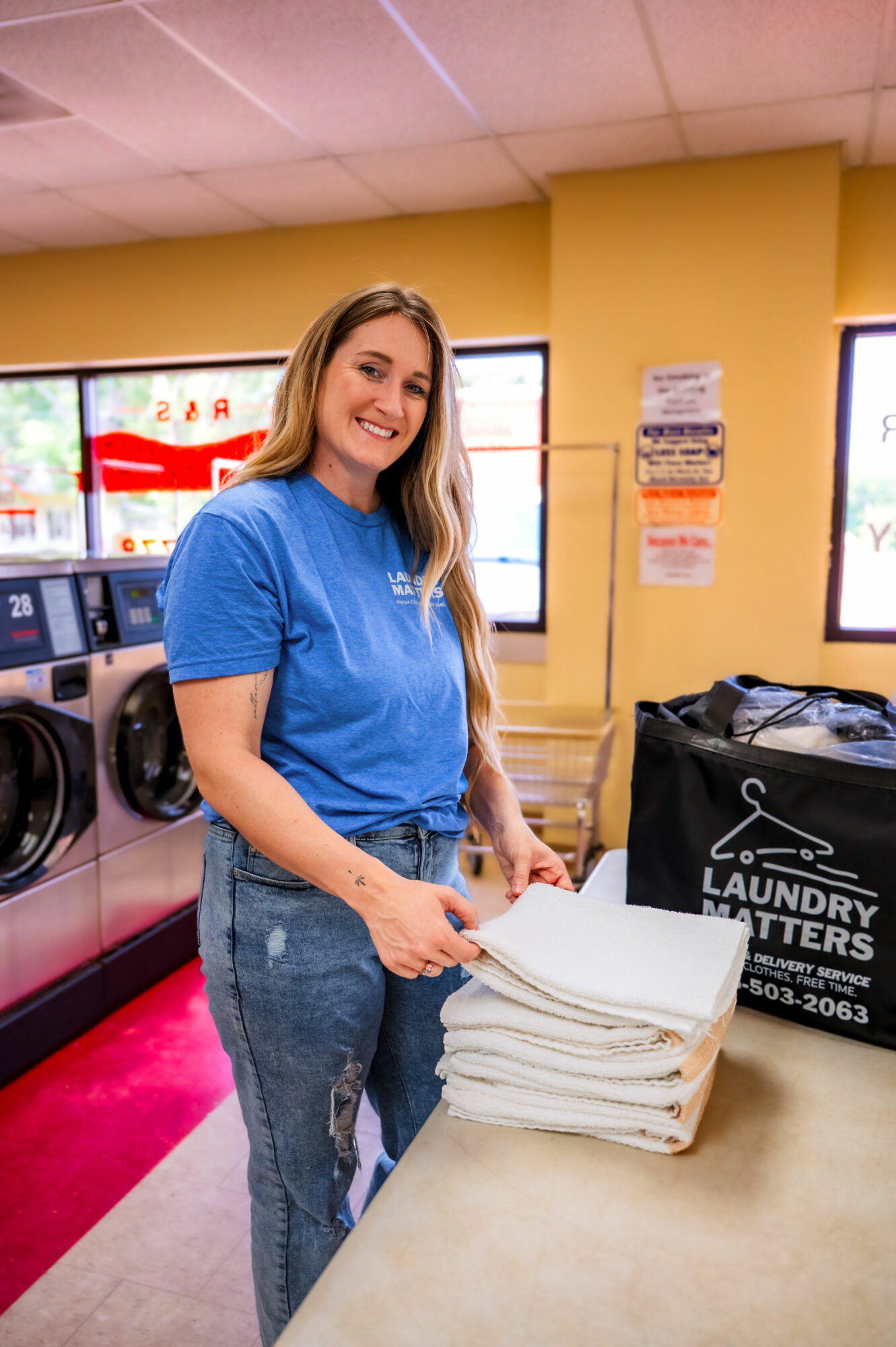 Woman in blue shirt folding laundry in laundromat, smiling, with washing machines and windows in background.