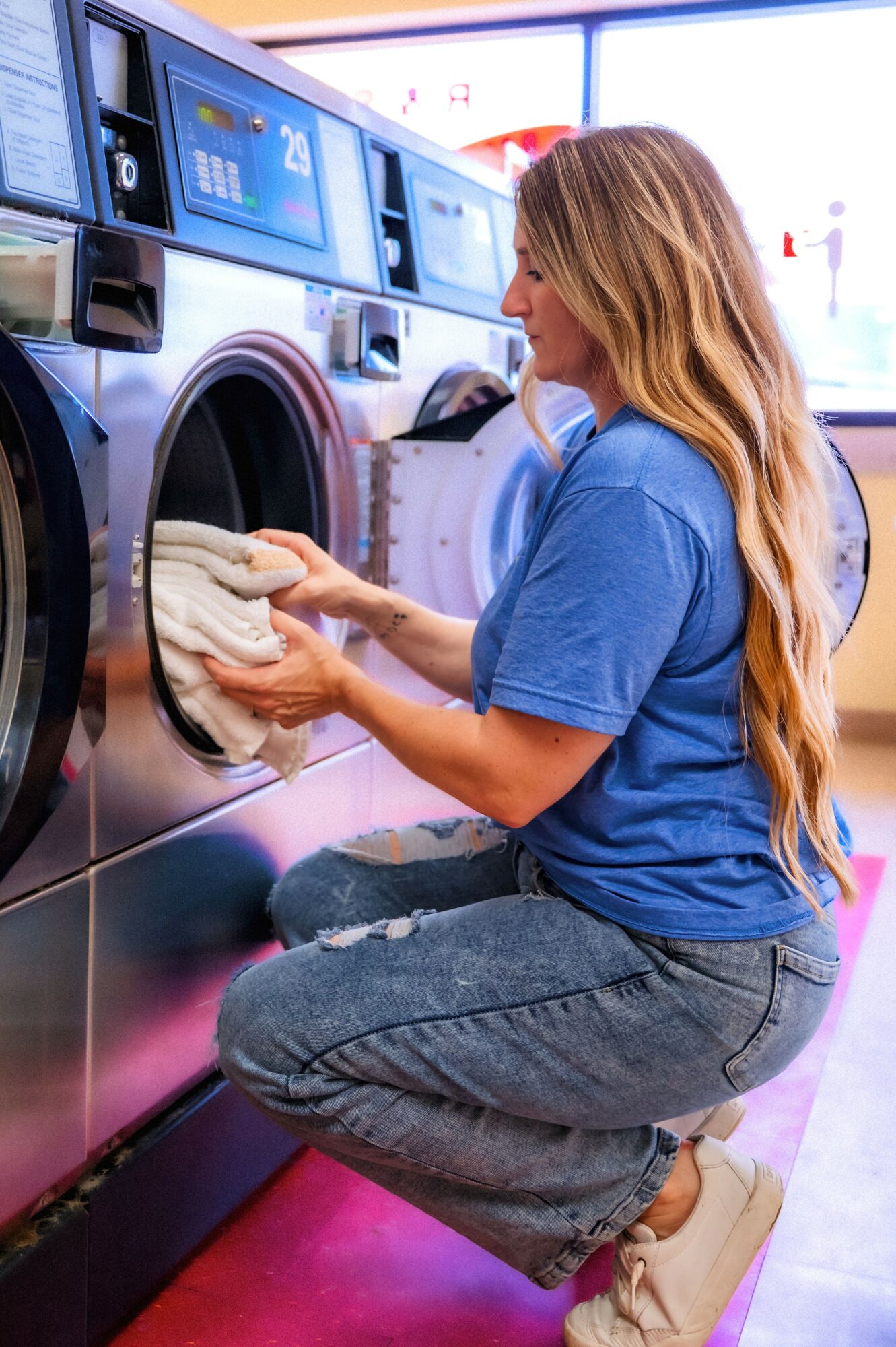 Woman loading laundry into washing machine, wearing blue shirt and jeans, in laundromat with large windows.