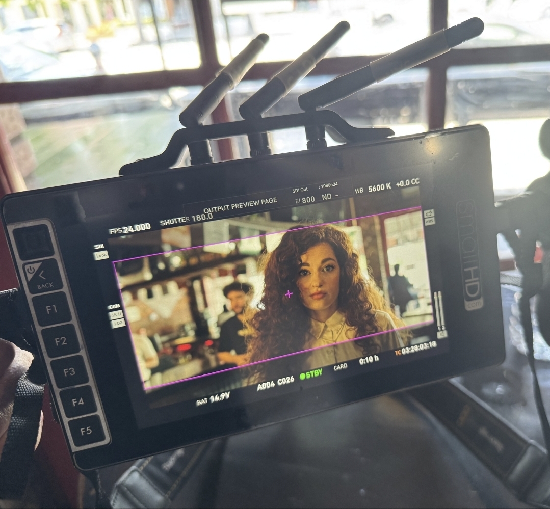 Camera monitor displaying a woman with curly hair in a public space, with people and windows in the background.
