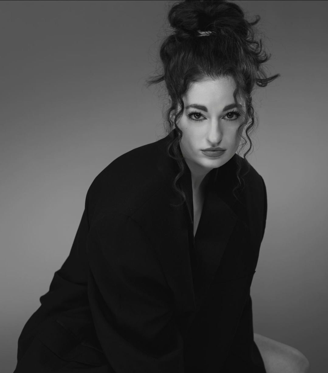 Woman with curly hair in a high bun, wearing dark clothing, looking at the camera against a plain background.