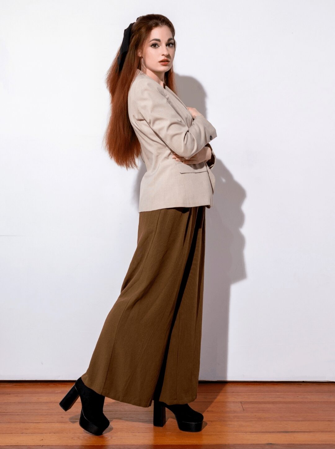 Woman with long hair in beige blazer and wide brown pants, standing sideways with arms crossed, against a white background.