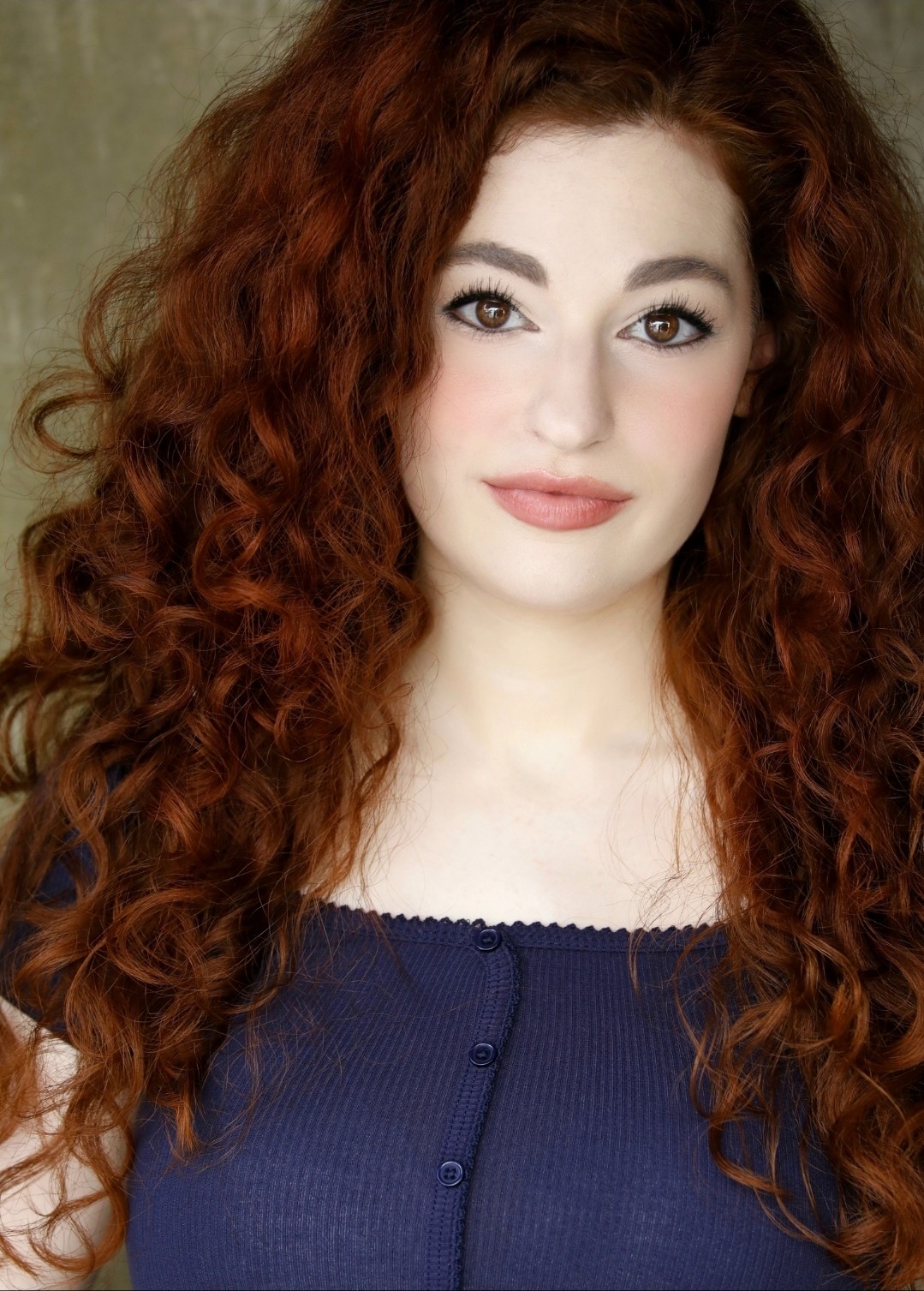 Young woman with long, curly red hair, fair skin, and wearing a dark blue top, smiling softly.