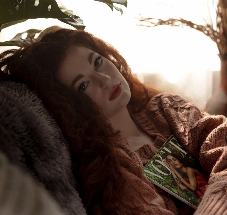 Woman with long curly hair lying on a couch, holding a magazine, with sunlight behind her.