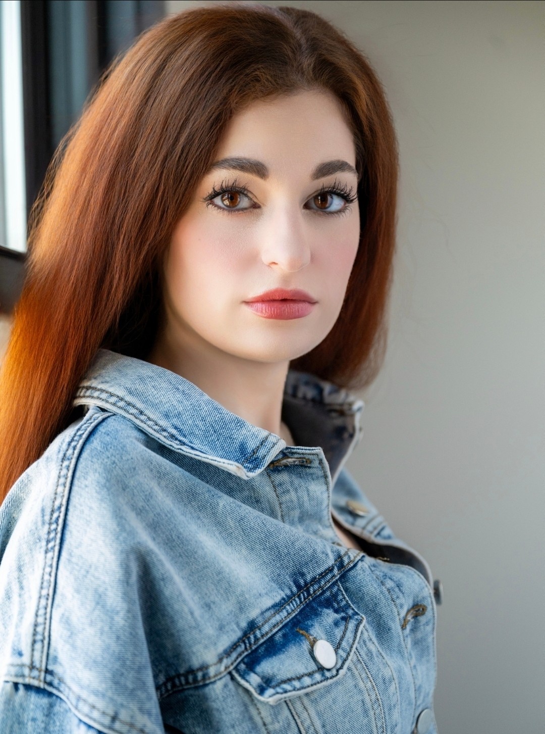 Young woman with long, reddish-brown hair wearing a denim jacket, looking at the camera with neutral expression.