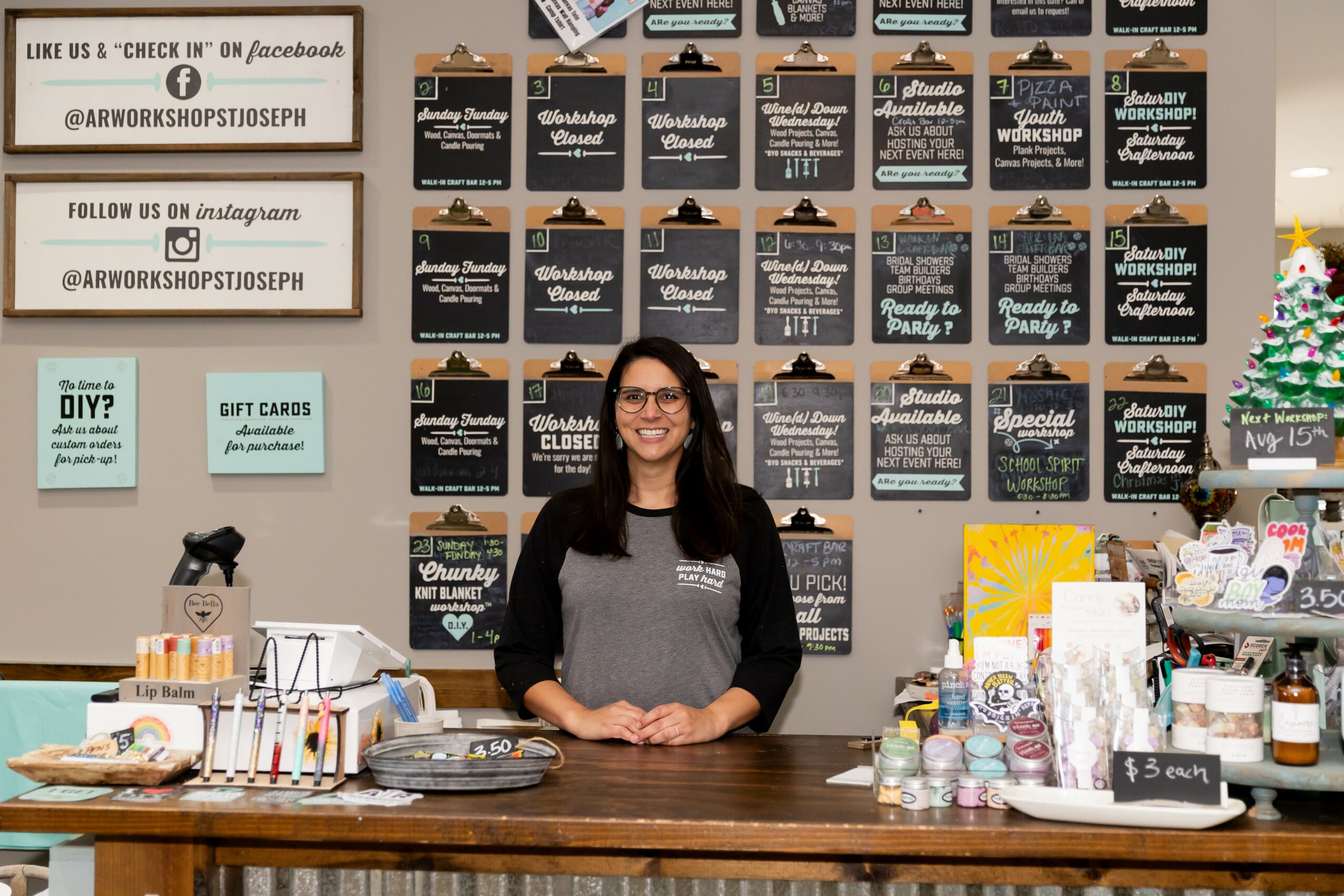 Woman with glasses standing behind a wooden counter with various craft supplies, smiling, in front of a black wall with signs and decorations.