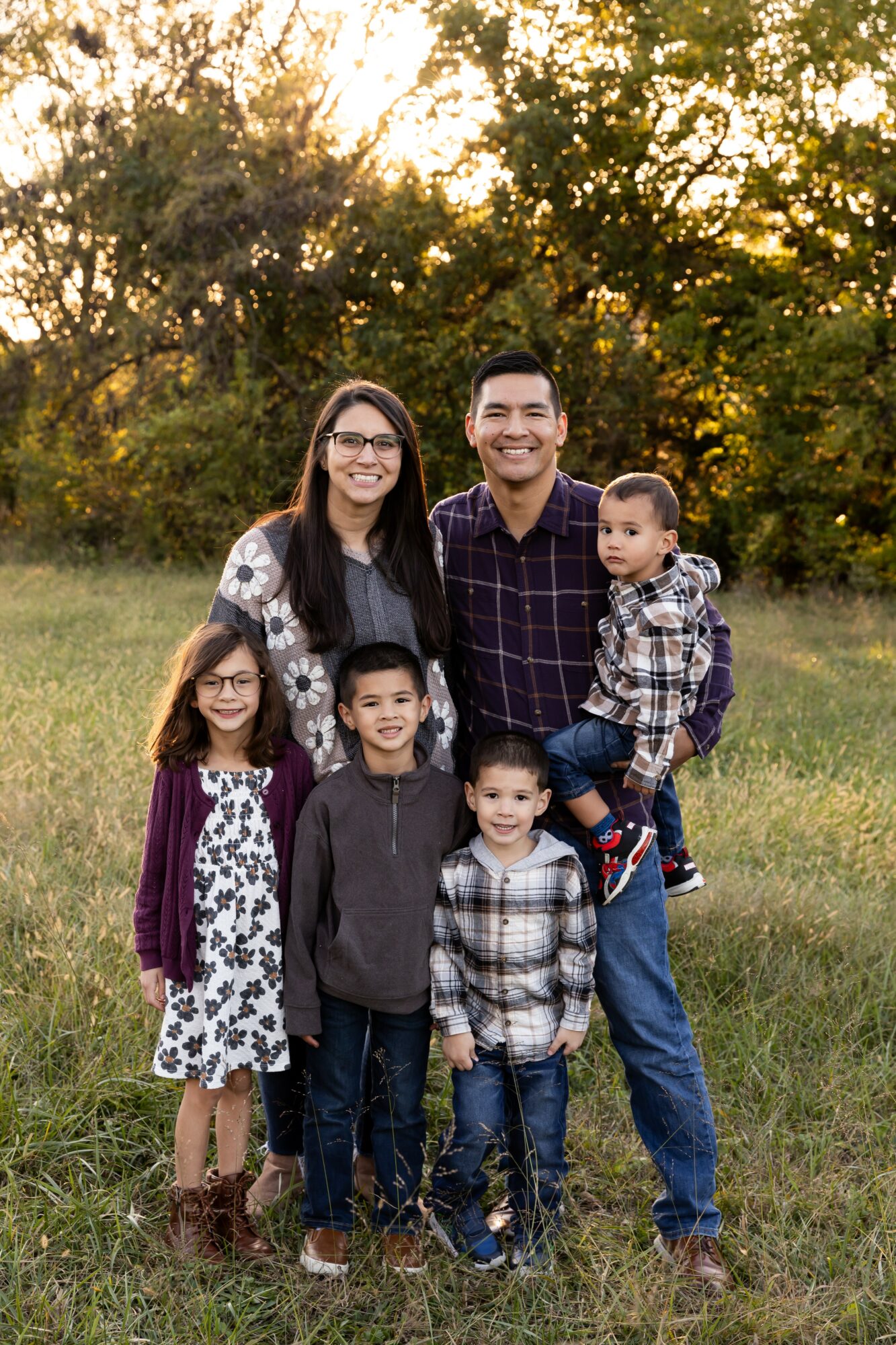 Family of six outdoors in a grassy area with trees, smiling, with adults and children, during daytime.