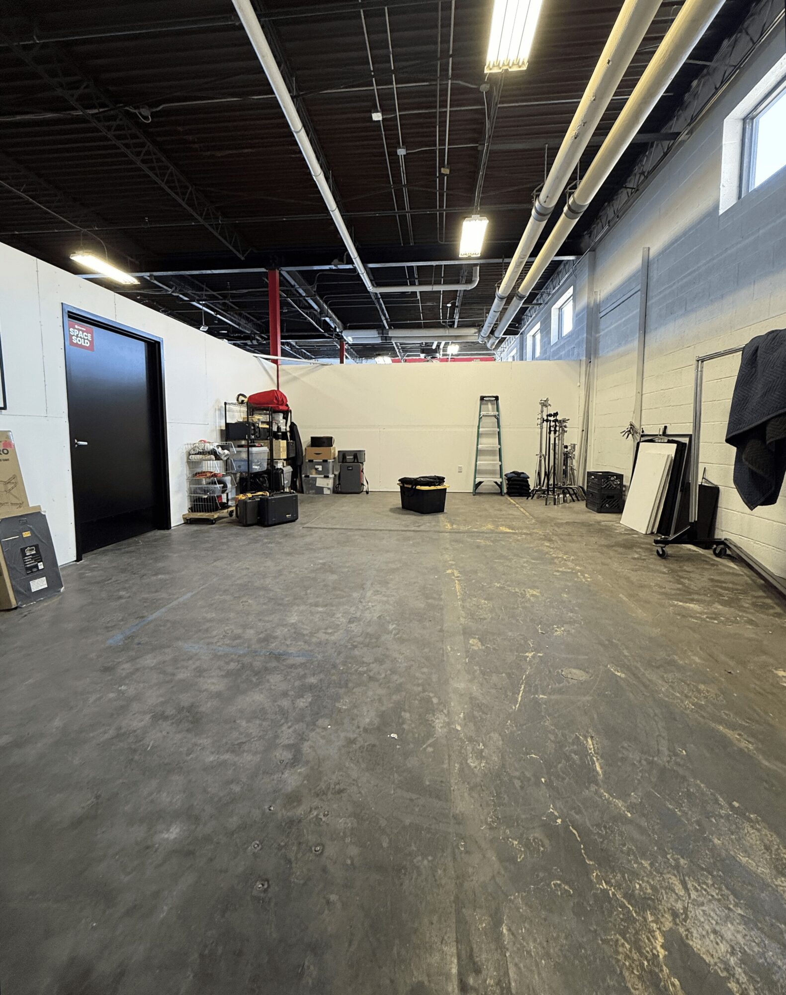 Industrial space with a concrete floor, black ceiling with pipes and lights, and a white wall with equipment and a ladder.