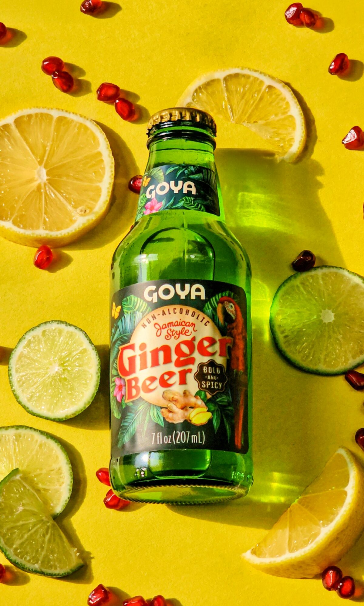 Goya Ginger Beer bottle surrounded by lemon, lime, and pomegranate seeds on yellow background.