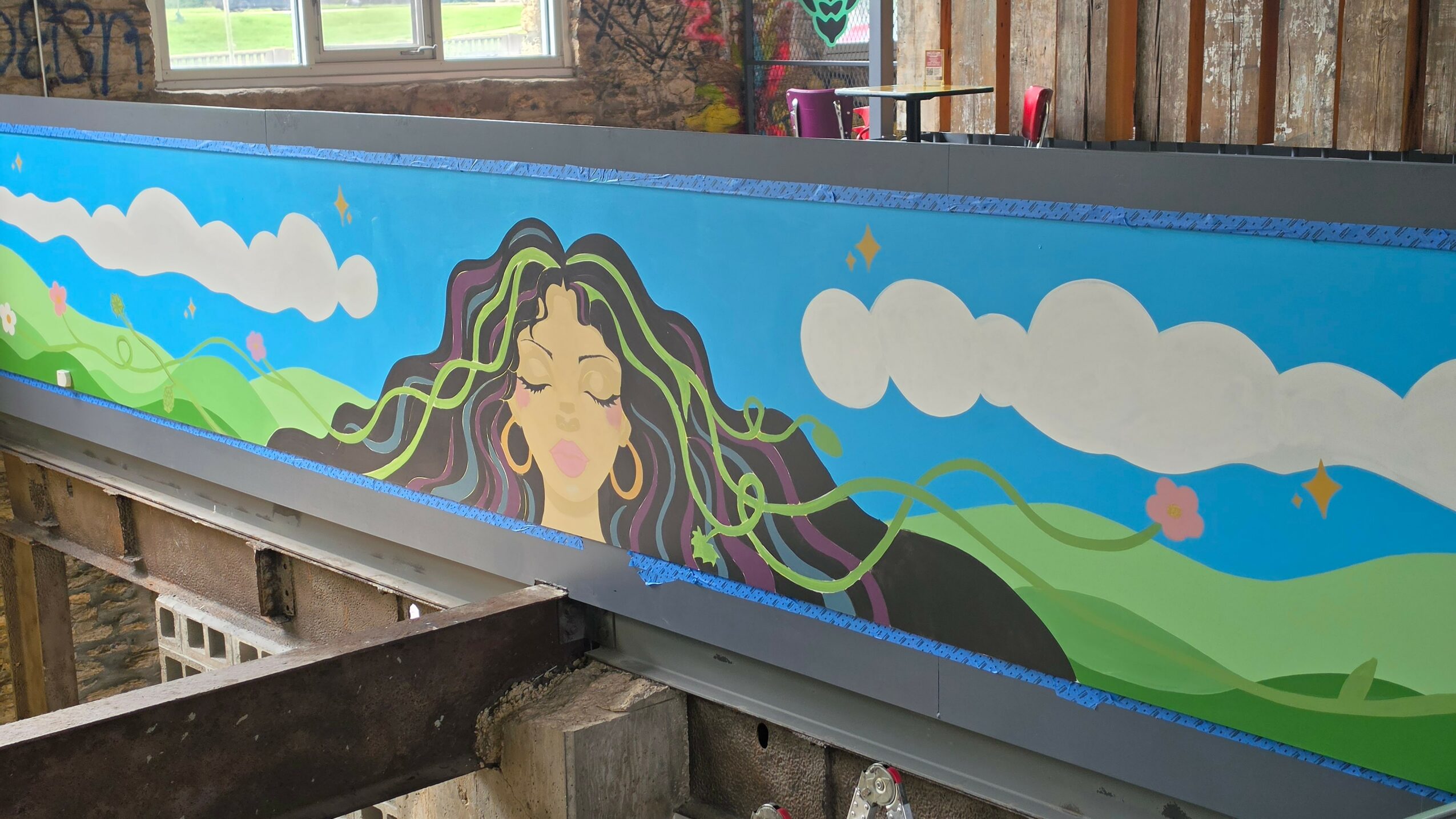 Colorful mural of a woman with flowing hair, green hills, blue sky, and clouds, painted on a long horizontal surface.
