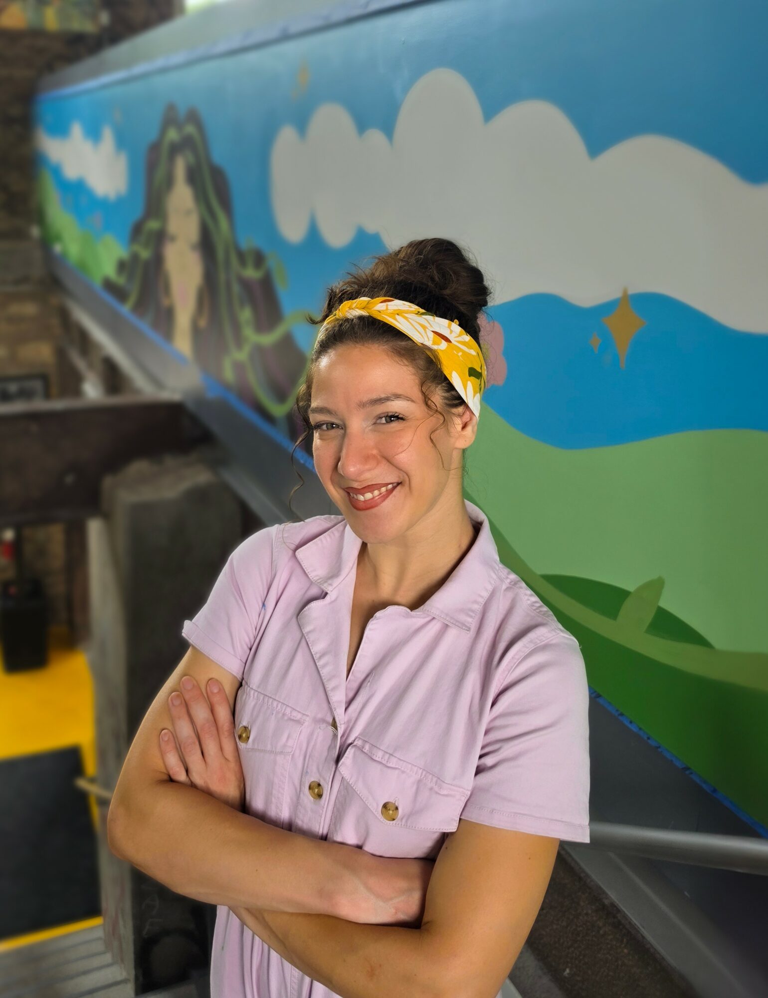Young woman with a yellow headband smiling, arms crossed, standing in front of a colorful mural with clouds and landscape.