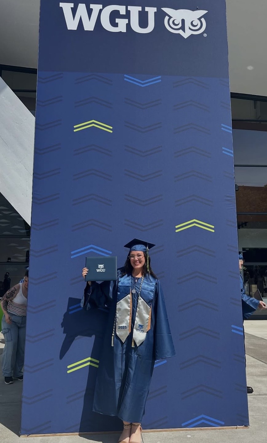 Person in graduation gown and cap standing in front of a large blue WGU banner with an owl logo, outdoors.