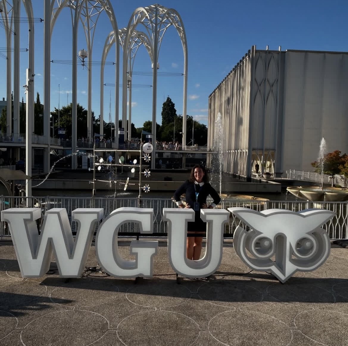 Person standing behind large letters spelling WGU with owl logo, outdoor setting, modern structures, clear sky.
