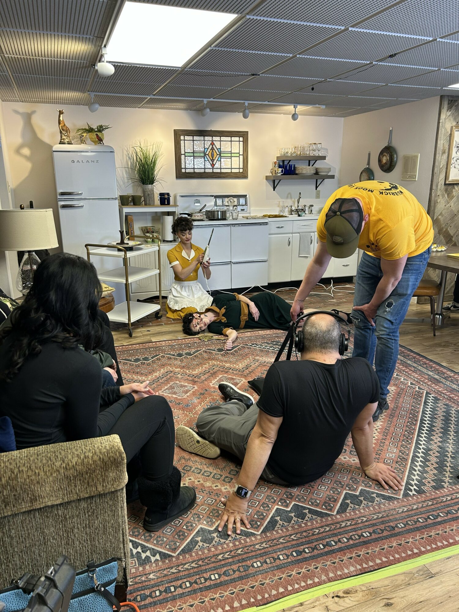 People in a room with a kitchen, some sitting on the floor, one lying down, and a person standing with a camera.