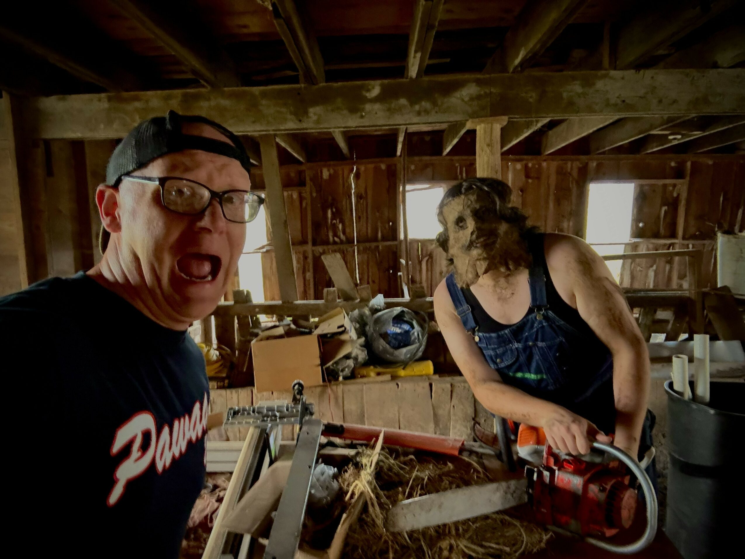 Man wearing glasses and a cap with a surprised expression, woman with curly hair and a mask, in a wooden workshop.