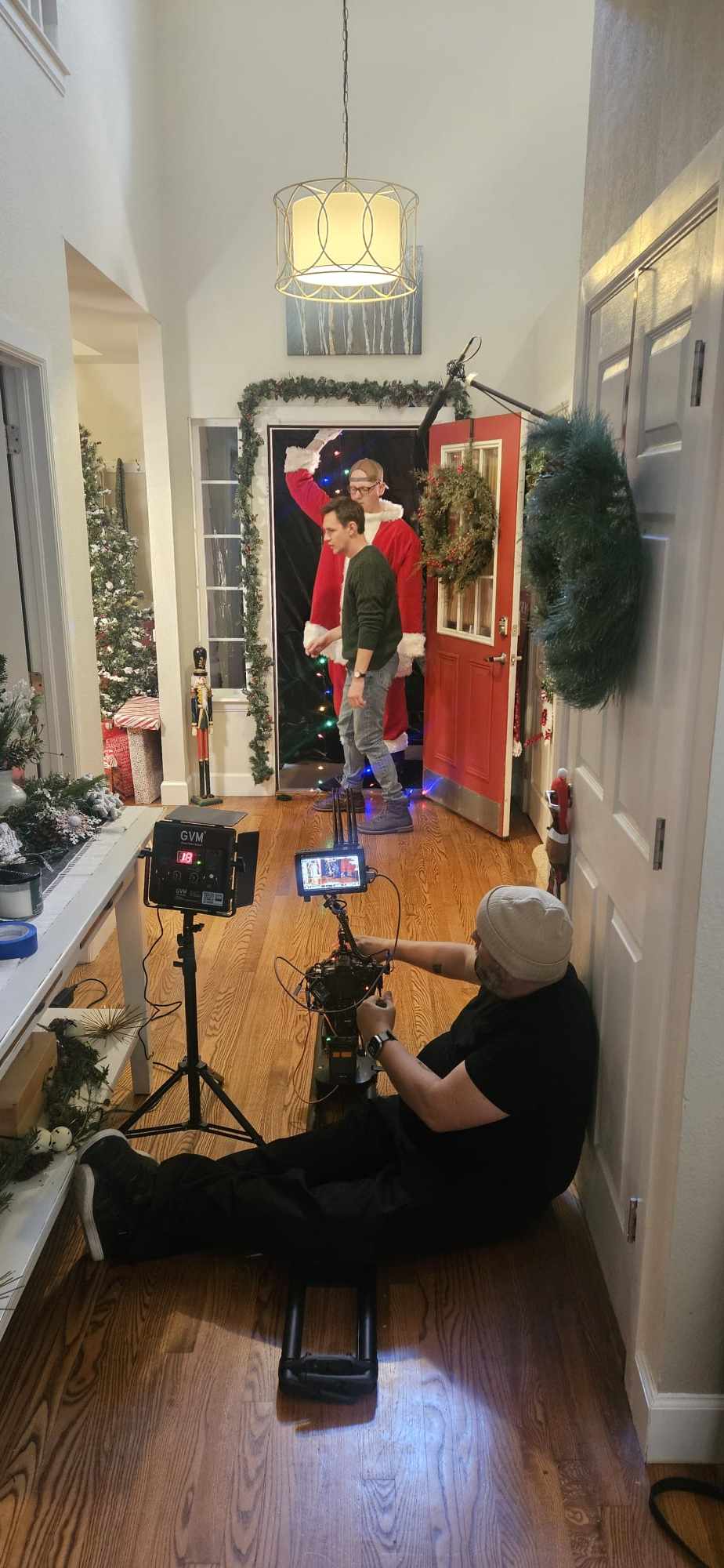 Person sitting on the floor with camera, filming a holiday scene with two people in front of a decorated door, Christmas wreaths, and lights.