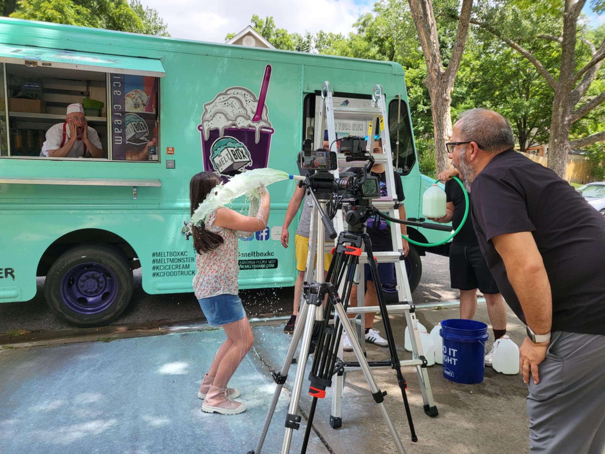 Child rinsing hair with water from a hose, person operating camera equipment nearby, ice cream truck in background, outdoor park setting