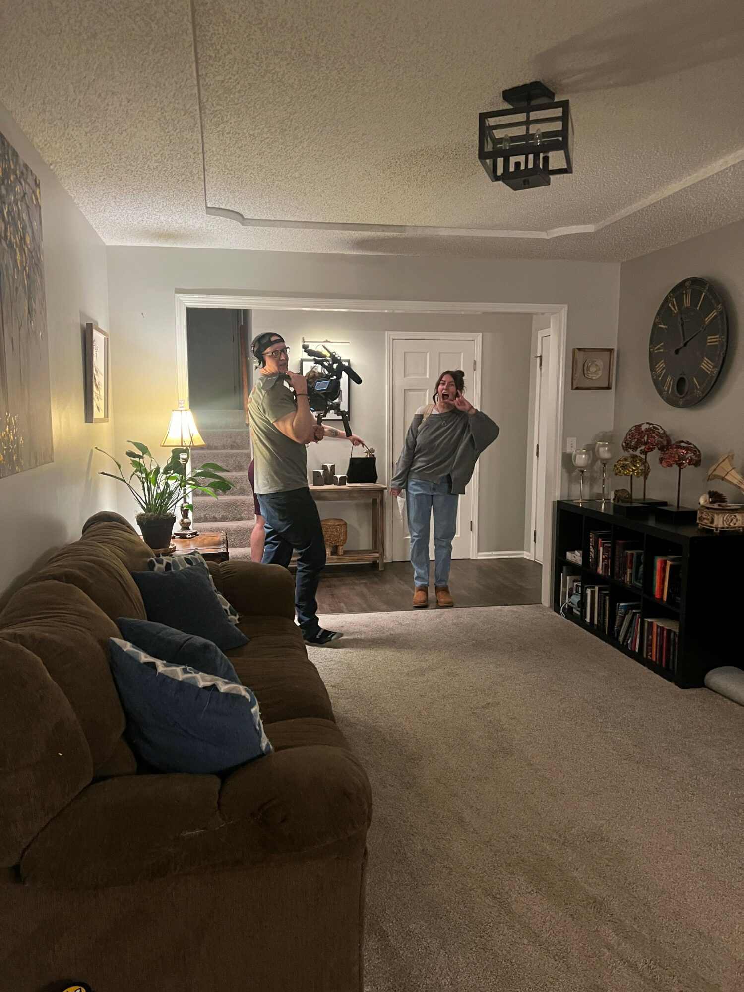 Person standing in doorway with a camera, two people in the background, living room with couch and bookshelf.