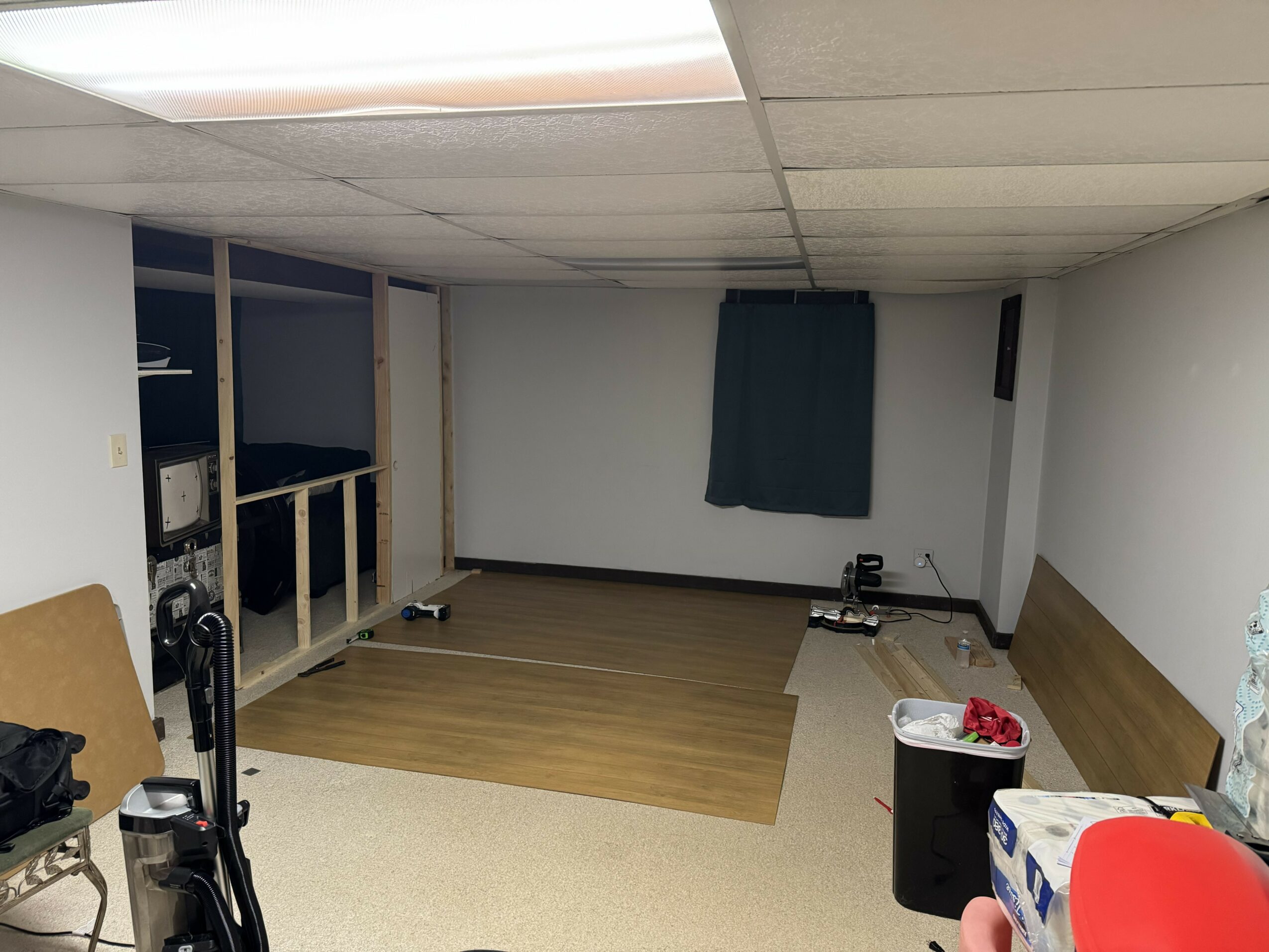 Room with a partially finished wall, a window with a curtain, and a mix of flooring types. Items include a trash bin and cleaning supplies.