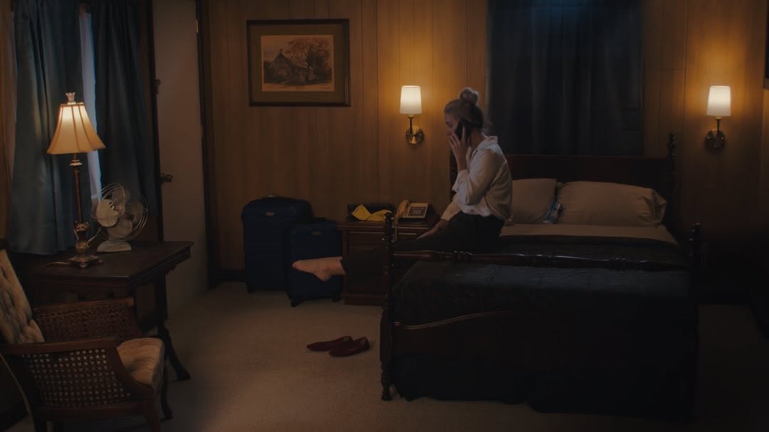 Person sitting on bed in dimly lit hotel room with nightstand, lamps, and window with curtains.