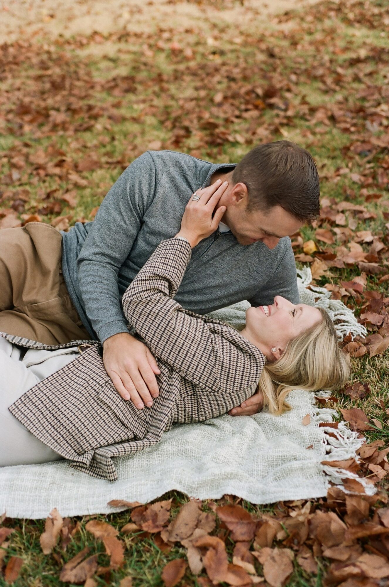 A couple lying on a blanket outdoors surrounded by fallen leaves, looking at each other closely.