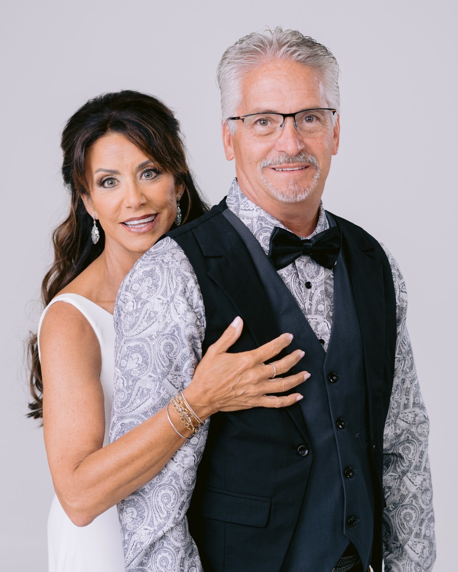 A smiling woman with dark hair and a man with gray hair and glasses pose together, dressed formally, against a plain background.