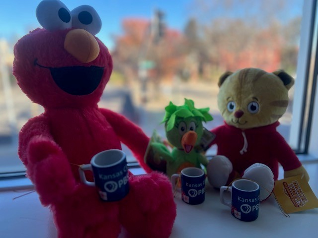 Elmo, a green plush toy, and a yellow plush toy with a red hoodie sit on a windowsill with cups, against a blurred outdoor background.